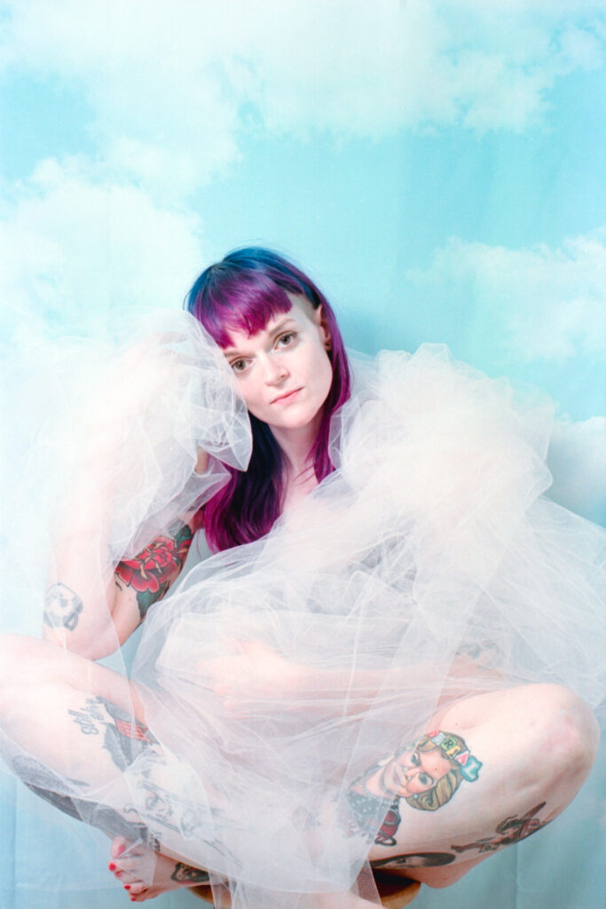 Portrait of photographer Amanda Wood dressed in a drape of crinkled blush tulle against a backdrop of cloudy skies