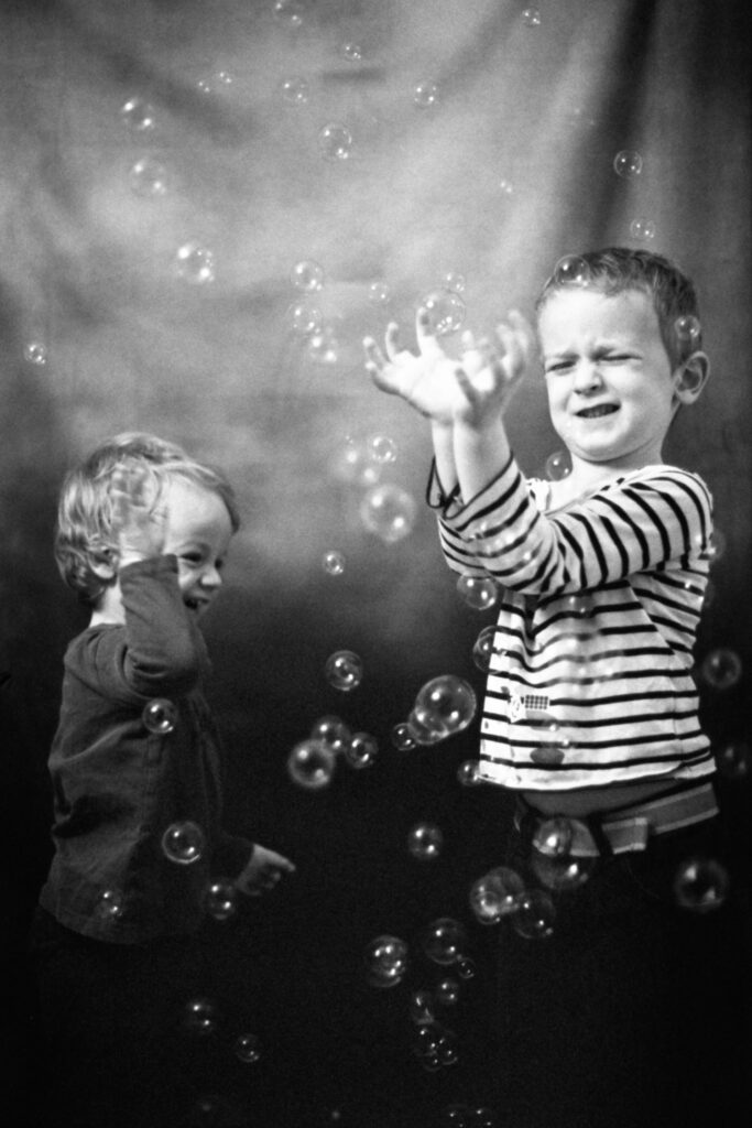 Monochromatic photograph of two young children playing with bubbles in a studio against a grey backdrop