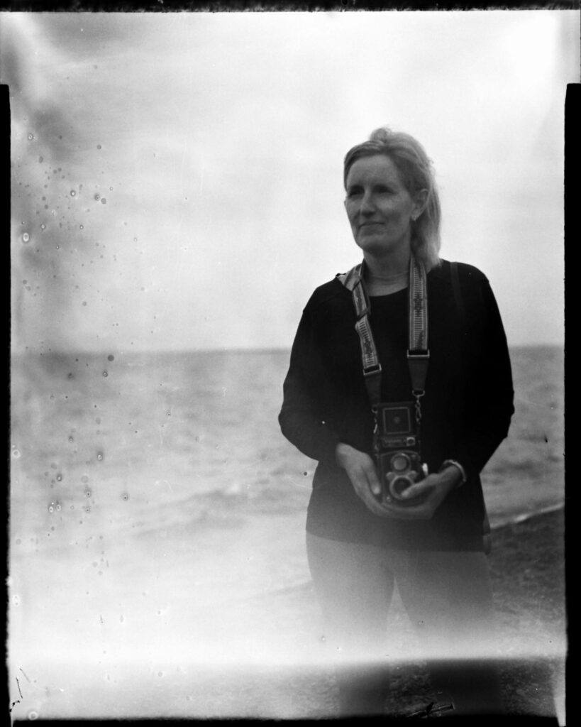 Large format black-and-white portrait of a woman holding a vintage twin-lens-reflex camera and looking into the horizon. The photograph has light leaks and small bubbles on the side due to alternative development techniques, giving it an out-of-time feeling.