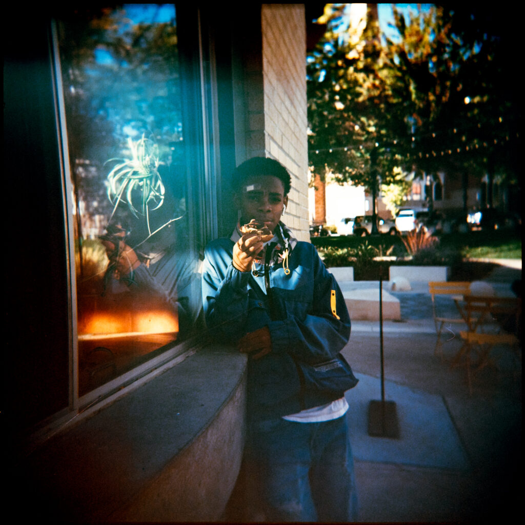 Portrait of a young black man with a bandaid on his forehead, eating a chocolate donut while leaning against a window in the public square