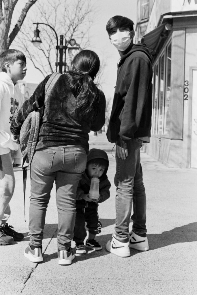Candid photograph of an asian-american family with every member looking in different directions, and a small child holding a milk bottle peeking from behind his parents looking straight at the photographer