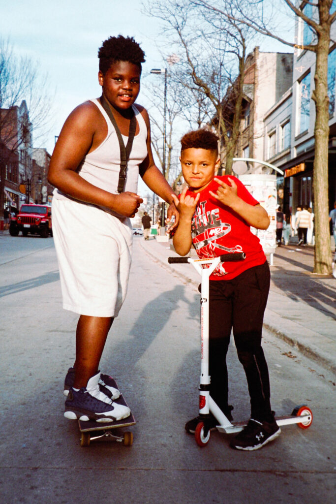 Two black kids posing for a photograph on their skate and scooter, in the golden light of a late spring afternoon