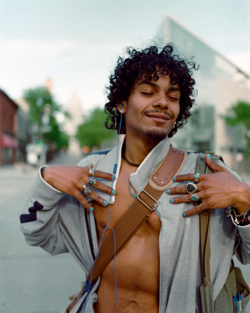 Portrait of a young man on state street with black curly hair and an open shirt, wearing finder jewelry and blue nail polish, feeling handsome and happy, looking like greek god Apollo