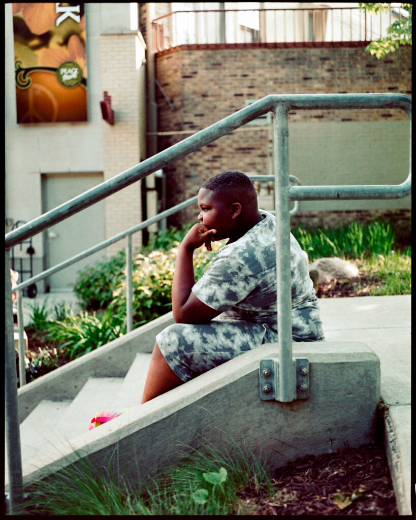 Candid profile portrait of a teenaged black boy, sitting with his chin resting in his hand in the manner of Rodin's famous statue "the thinker"