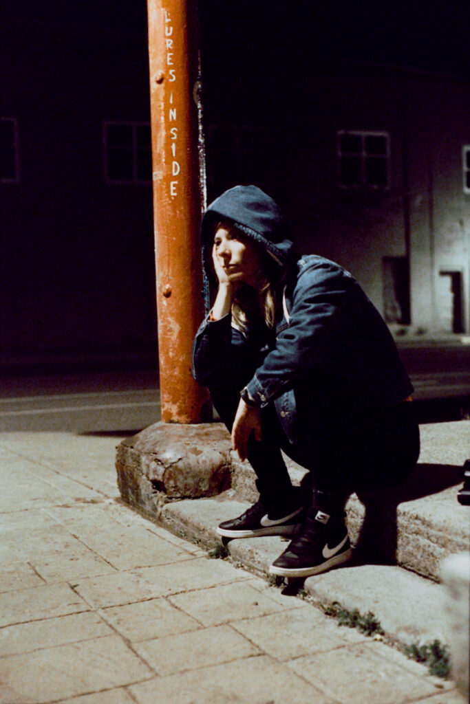 Night portrait of a woman sitting, looking bored, on a shop doorstep in a small, quiet town, with a sign saying "lures inside" over her head
