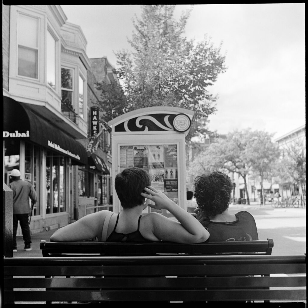 Black-and-white photograph of two people sitting on a bench, shot from behind them, with one of the subjects folding his arm behind the other subject's back in a trusting and comfortable gesture