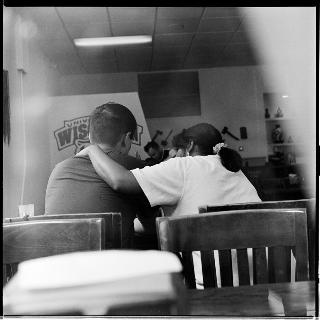 Black-and-white photograph taken from outside a window of two people with their back turned: a woman comforting a young man inside a restaurant on University of Wisconsin, Madison's campus