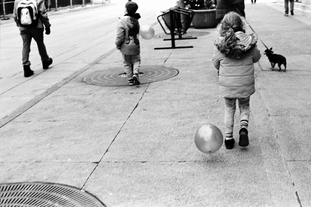 Black-and-white street photograph of two children walking with balloons