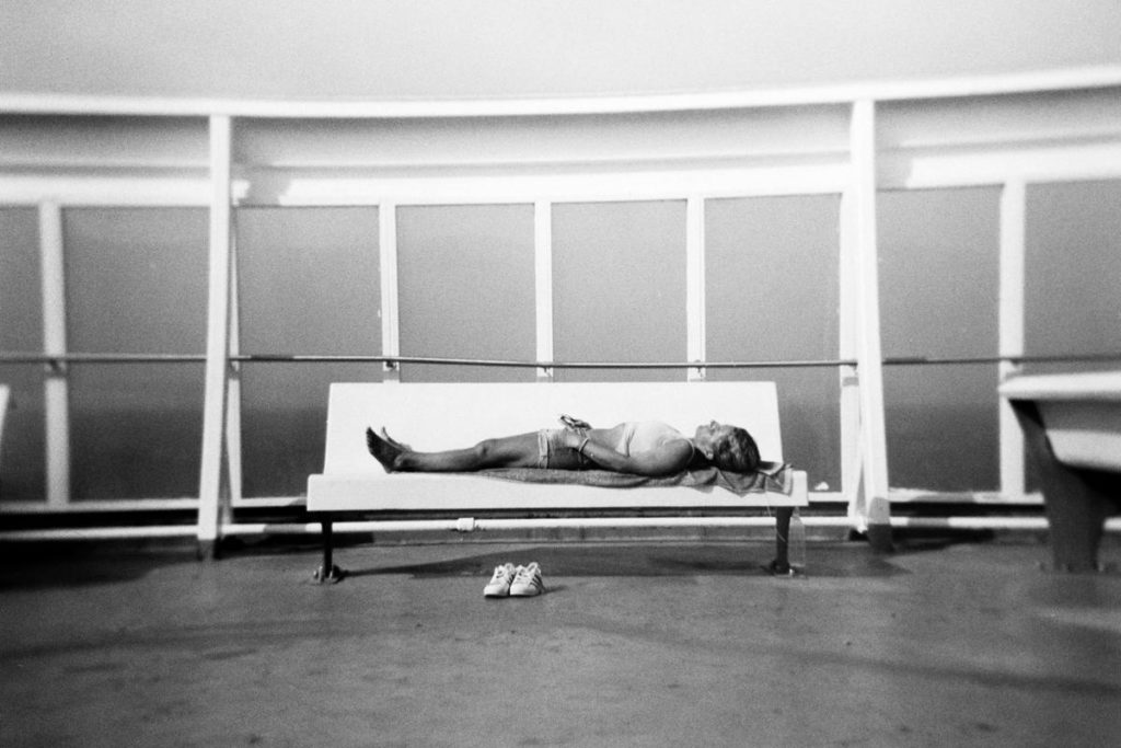 black and white street photograph of a young woman napping on a bench on the top deck of a ferry boat, with her shoes neatly left on the ground