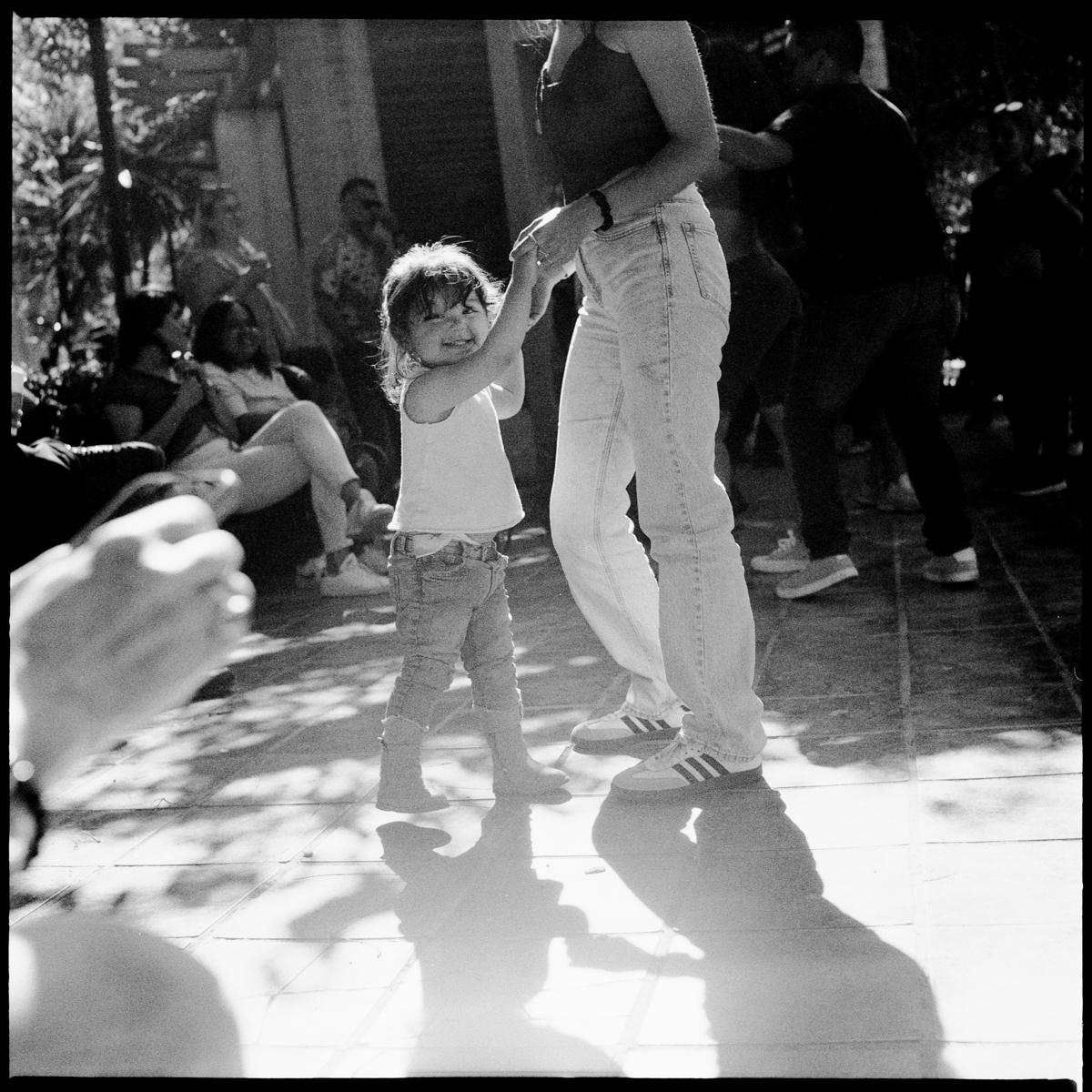 analog black-and-white street photograph of a kid dancing with her mom, in Parque México on a Saturday afternoon
