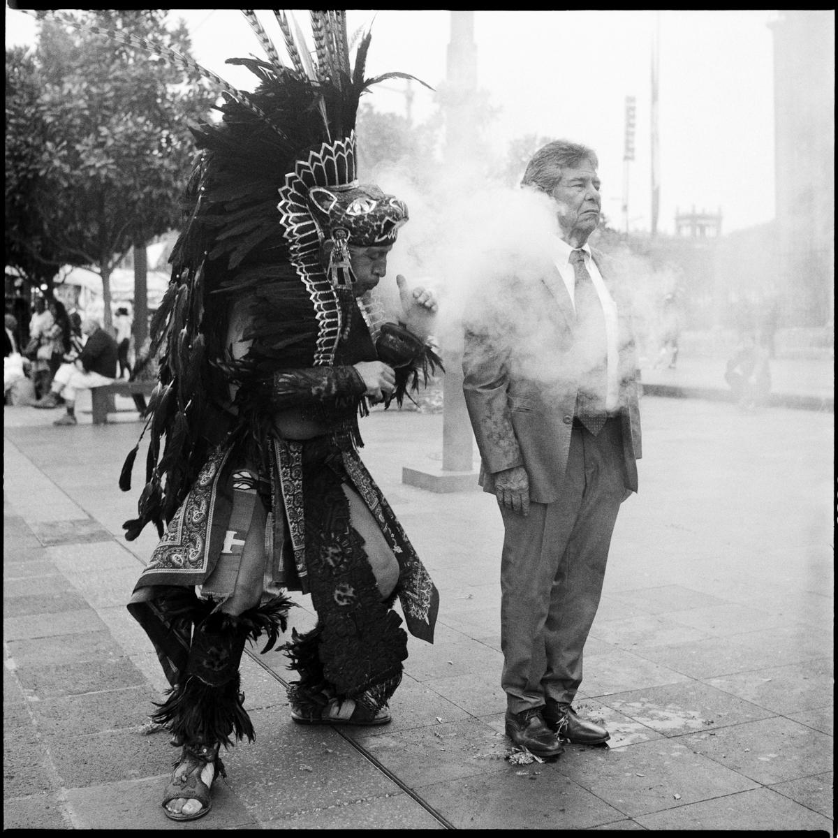 analog black-and-white street photograph of an elderly businessman getting an Aztec cleanse in Mexico City's city center