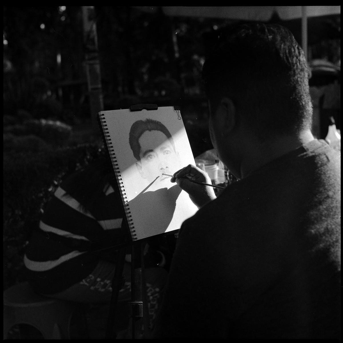 analog black-and-white street photograph of a drawing being drawn by the artist, in Parque México on a Saturday afternoon. The drawing is exposed properly, while everything else is in dark stark shadow.