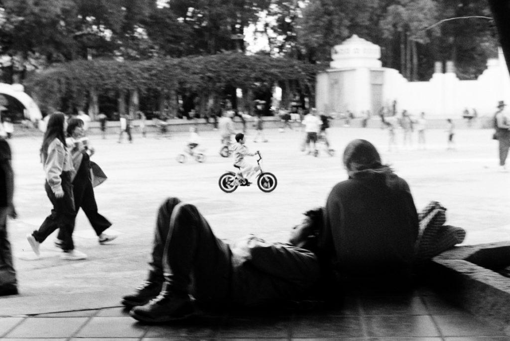 analog black-and-white street photograph of a kid on a bicycle, with a couple hugging in the foreground , in Parque México on a Saturday afternoon
