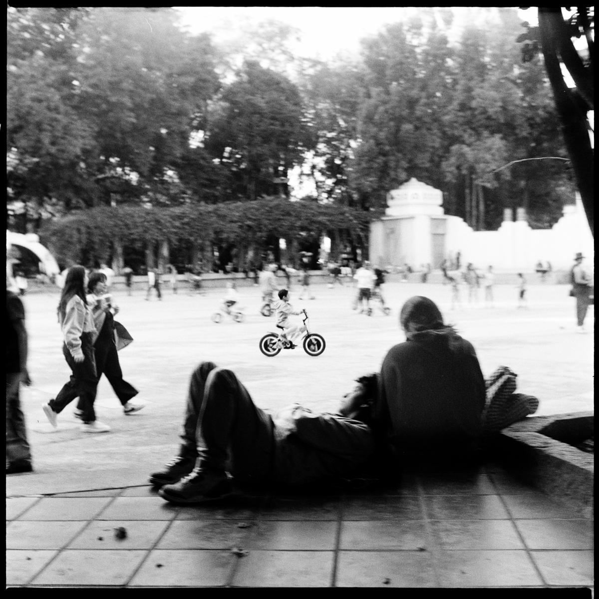 analog black-and-white street photograph of a kid on a bicycle, with a couple hugging in the foreground , in Parque México on a Saturday afternoon