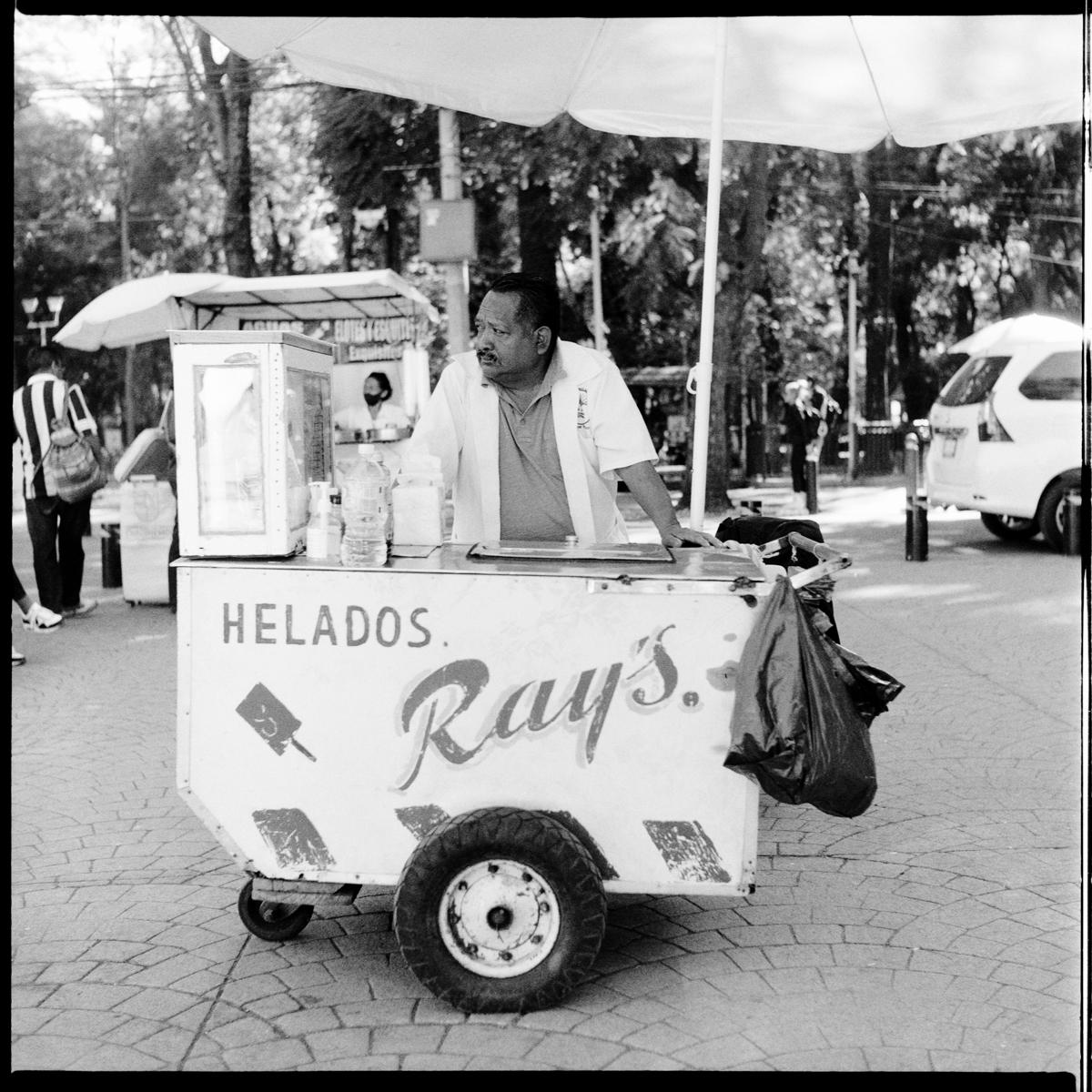 analog black-and-white street photograph of an ice-cream street food vendor, in Parque México on a Saturday afternoon
