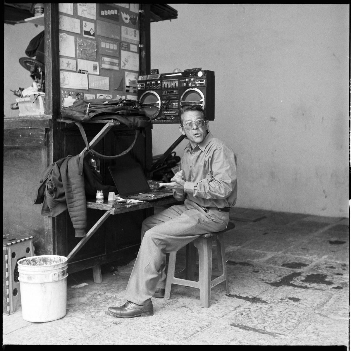 analog black-and-white street photograph of a man sitting at his street stand. He is a printer by a church in Mexico City's historic center, and there is an old boombox by his side.